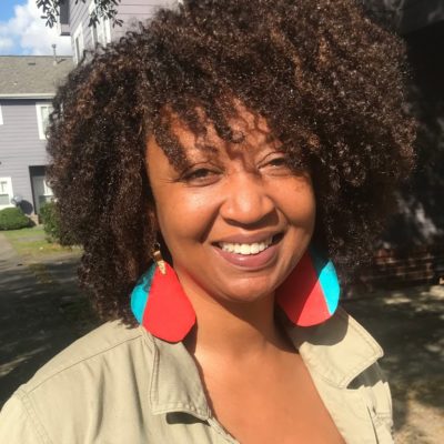 Headshot of Shondra Muhammad in a tan jacket with blue and red earrings