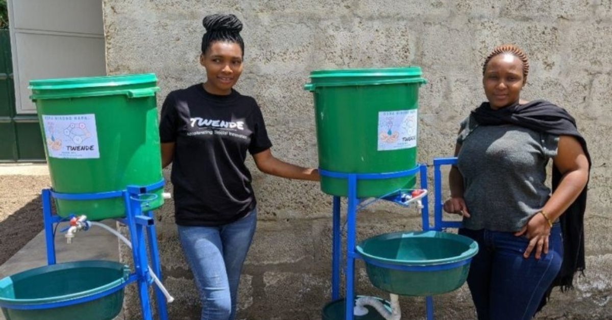Two women standing in front of a foot-operated hand-washing station. Innovative nonprofit solution