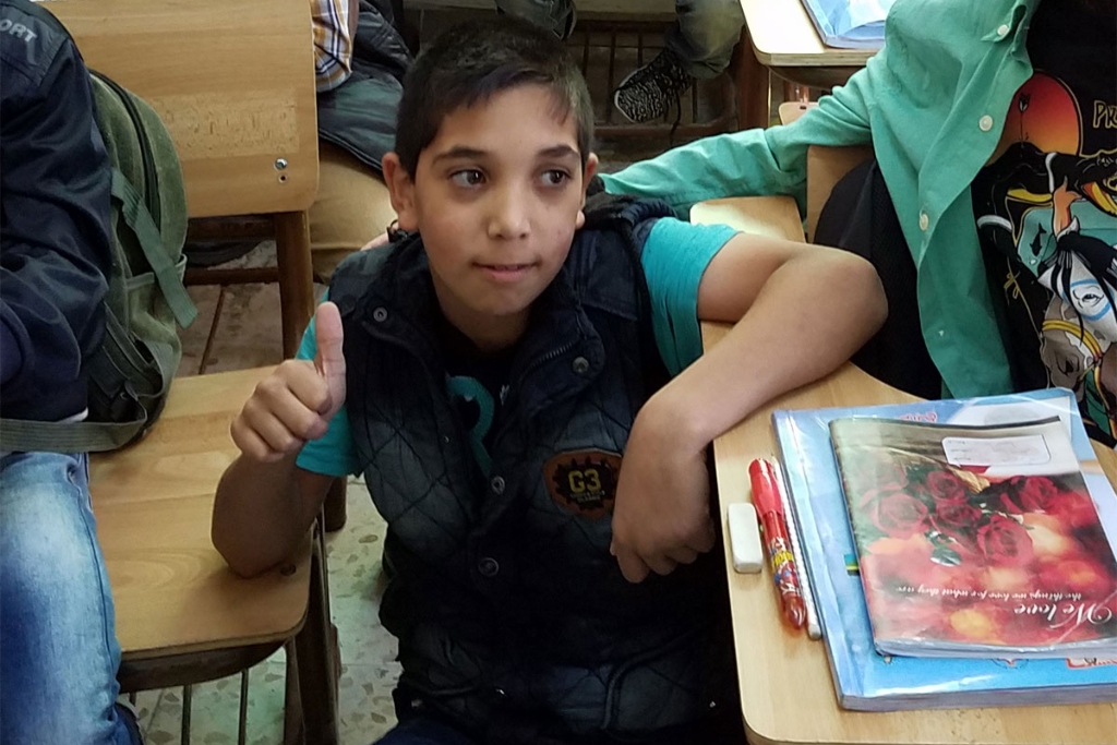 A young boy in a black vest sits at a school desk and puts his thumb up. Other students are at desks around him