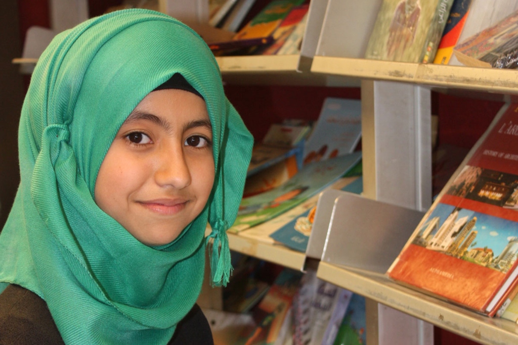 A young girl in a green head covering smiles. Shelves of books are behind her