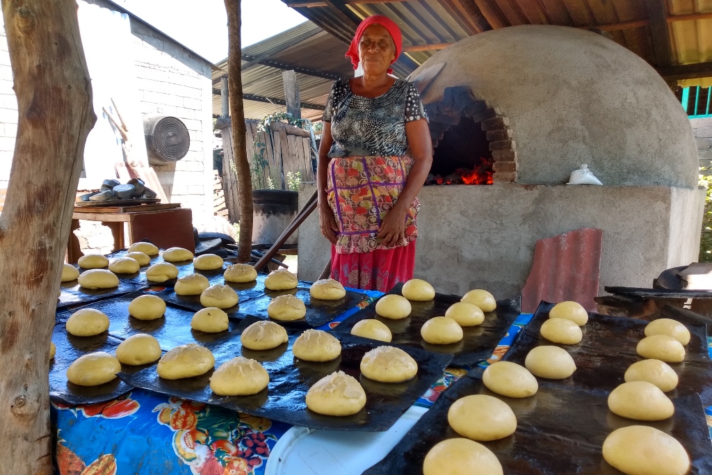 A woman stands behind sheets of unbaked buns. A large cement stove is in lit the background.