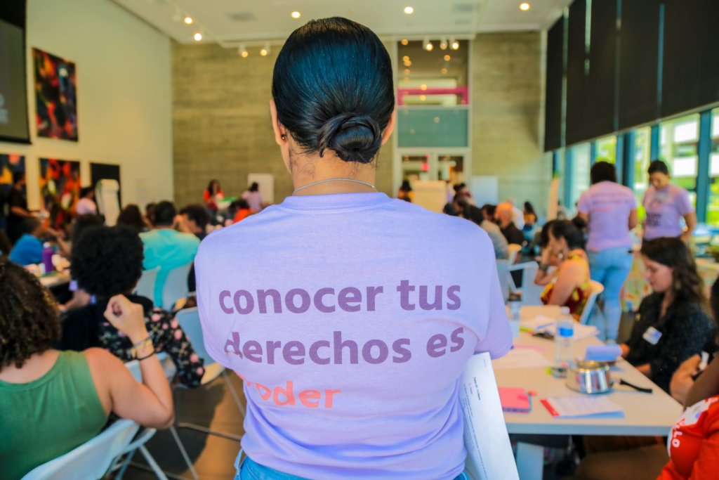 A woman in a purple t-shirt with the words "conocer tus derechos es poder" on the back stands in front people seated at tables.