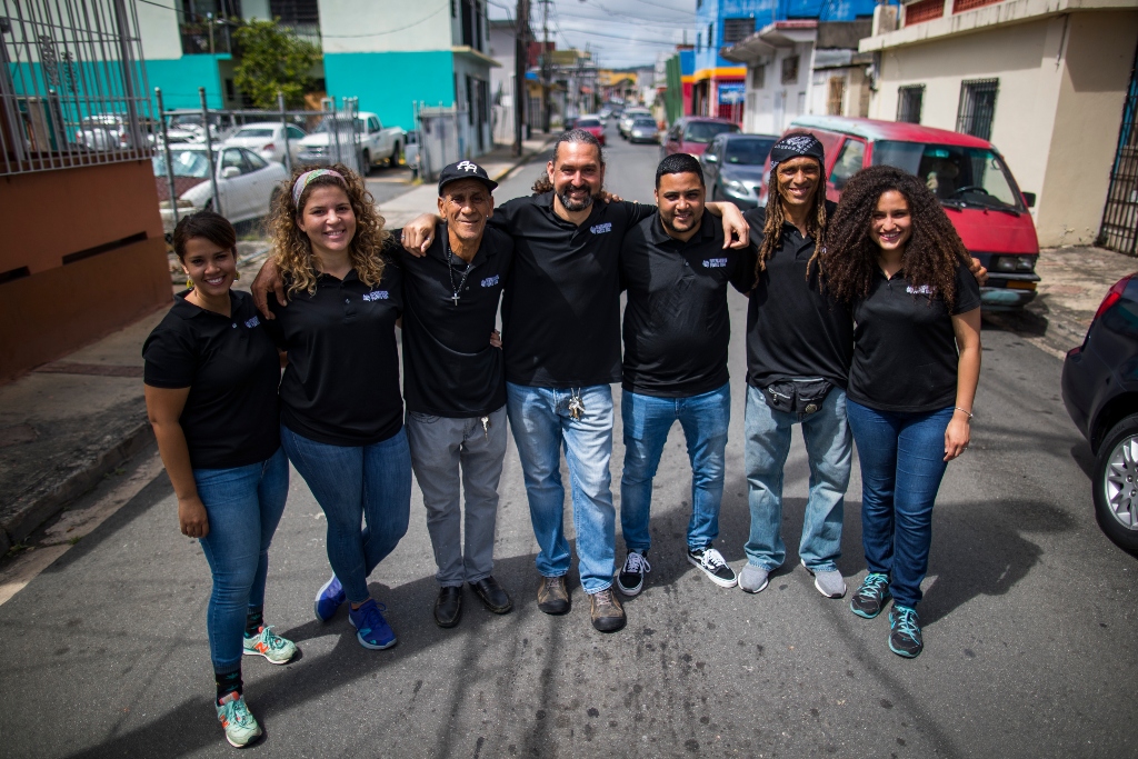 A group of people in matching black shirts stand in a group with their arms around each other in a Puerto Rican street.