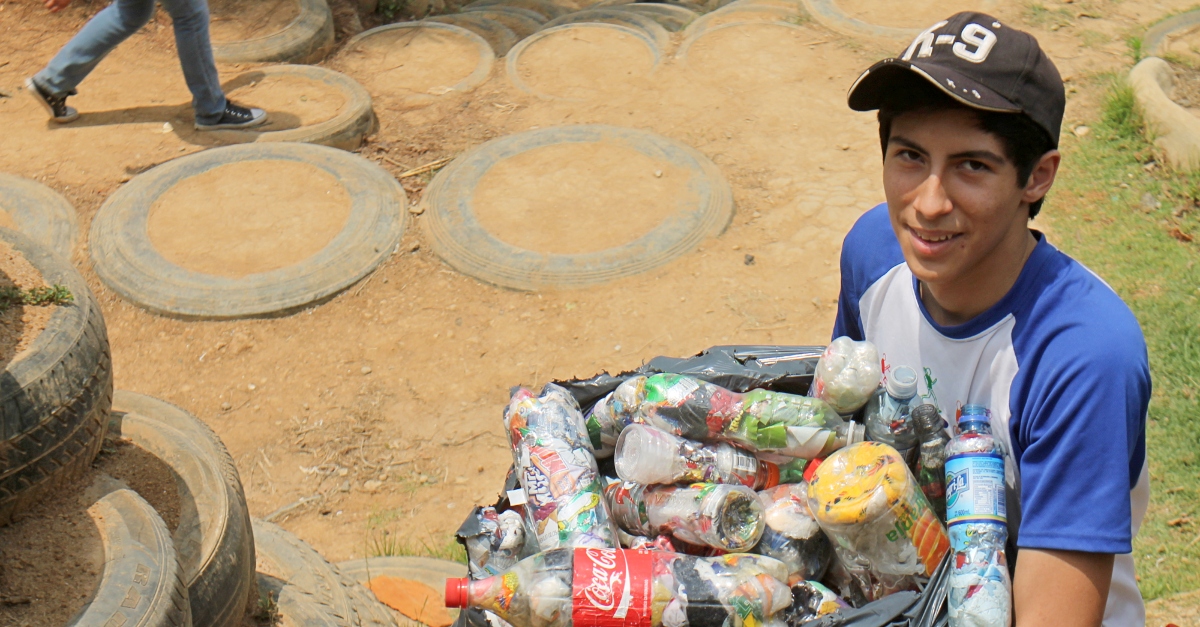 A young boy holds a bag of cans and bottles by a tire wall, part of an innovative education program teaching kids to transform trash into community infrastructure