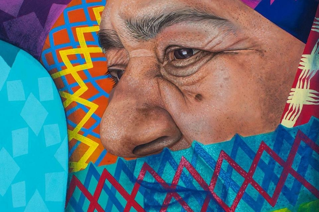 A closeup of a mural showing a woman's profile with her mouth covered by a bright blue and red pattern. Colorful patterns surround her face.
