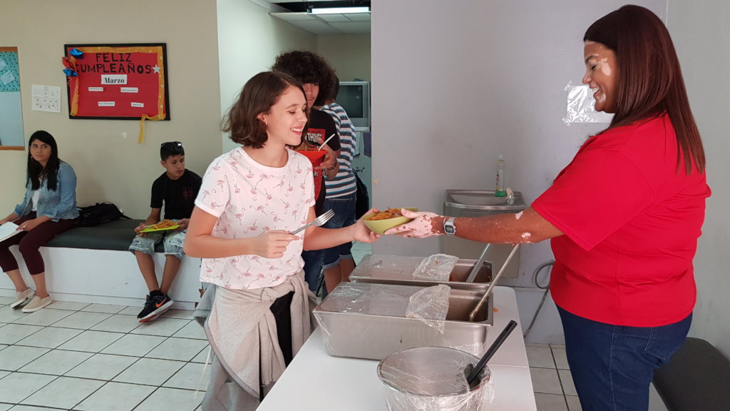 A woman serves a bowl of food to a student in a cafeteria. Another Student stands behind her in line.
