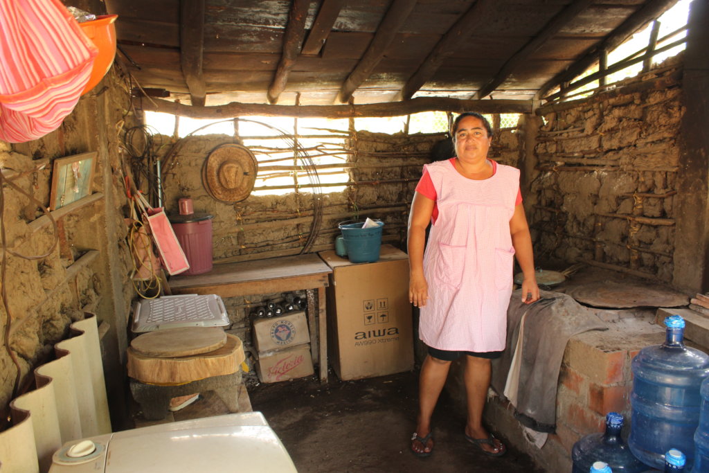 A woman in a pink apron stands in the corner of her kitchen in front of her reconstructed brick stove 