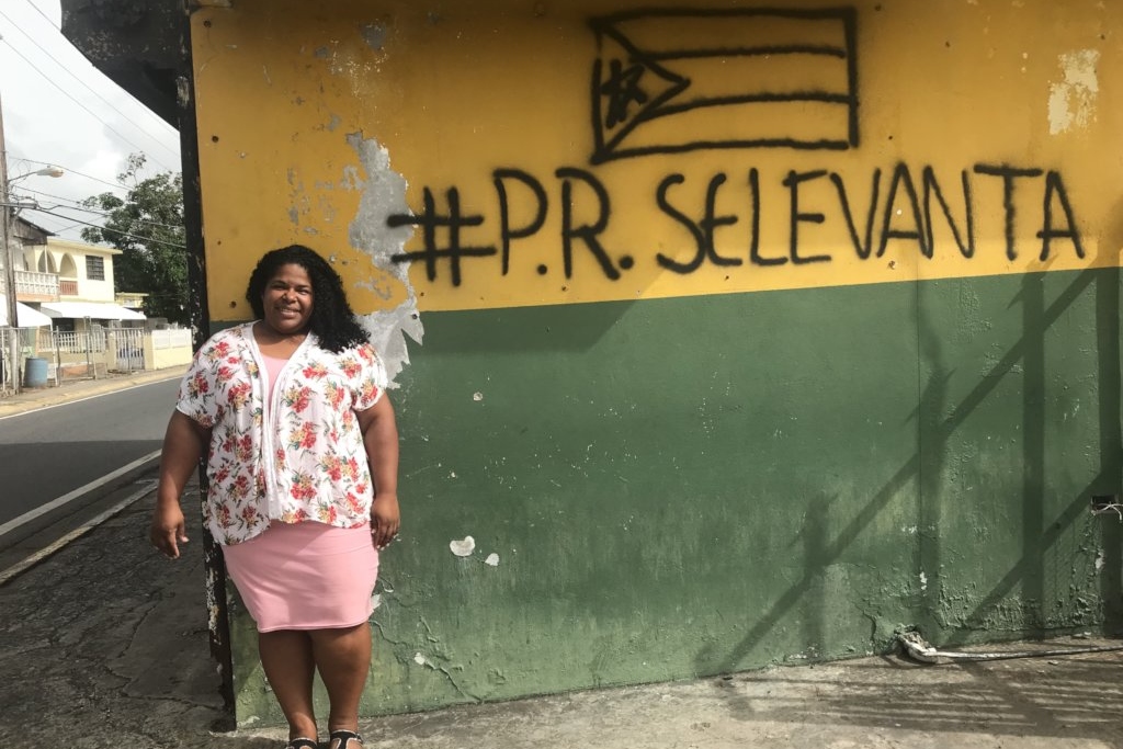 A woman in a pink flowered top and a darker pink dress stands in front of a wall painted yellow and green. The words #P.R. Selevanta and the Puerto Rican flag spray painted at the top of the wall.