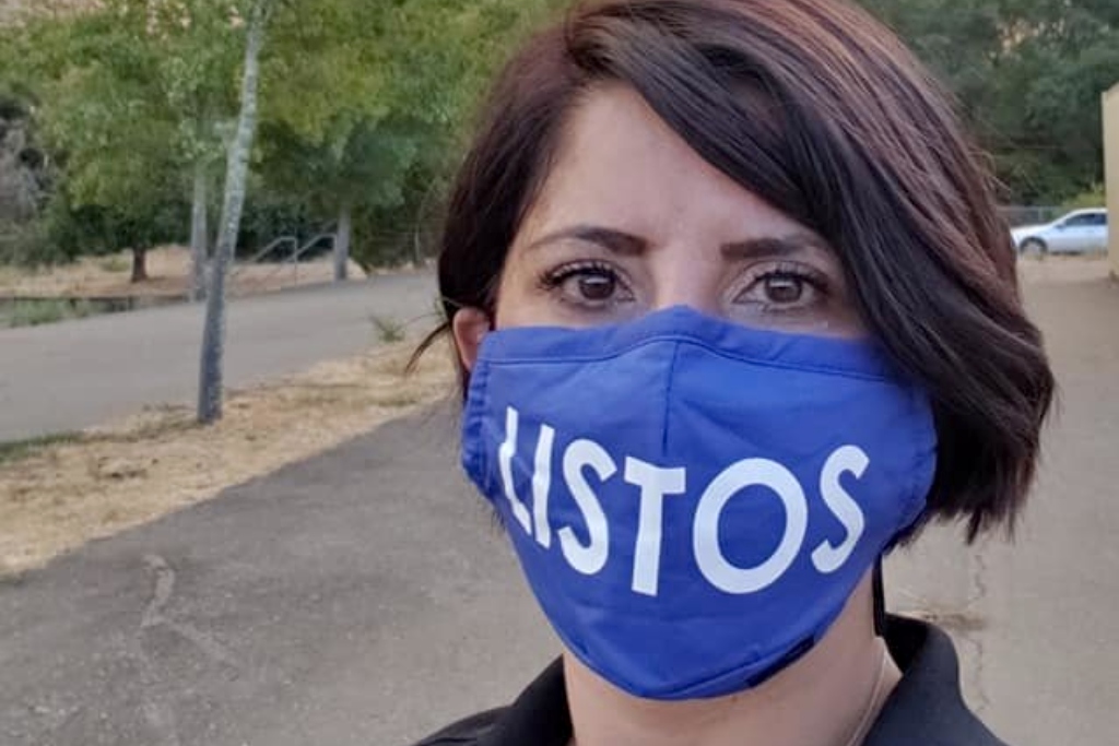 A woman wearing a blue mask with the word "Listos" stands in front of a smoke-filled sky,