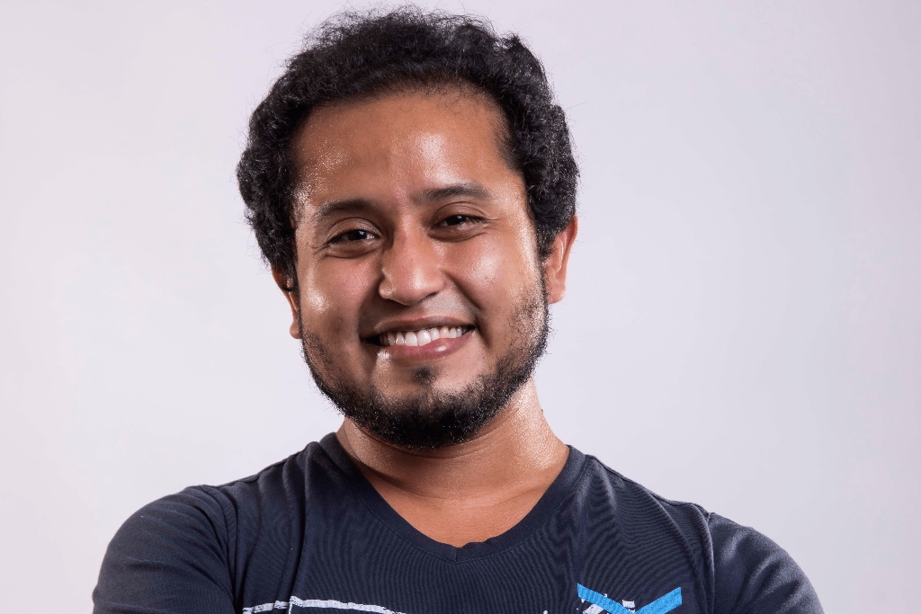 A man wearing a blue t-shirt smiles in front of a gray background