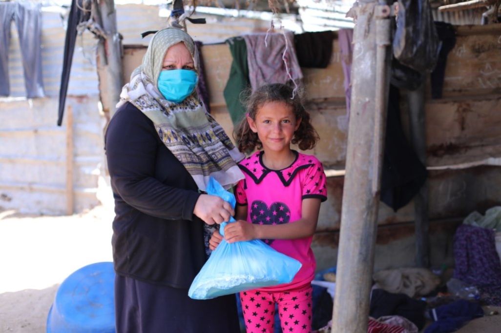 A woman wearing a blue mask and head covering hands a plastic bag containing food to a child wearing pink clothes.