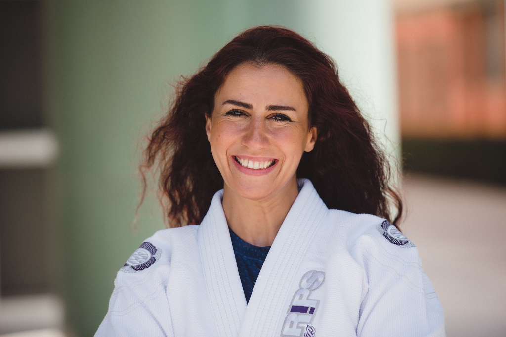 A woman wearing a white jiu jitsu robe stands smiling in front of a light green background