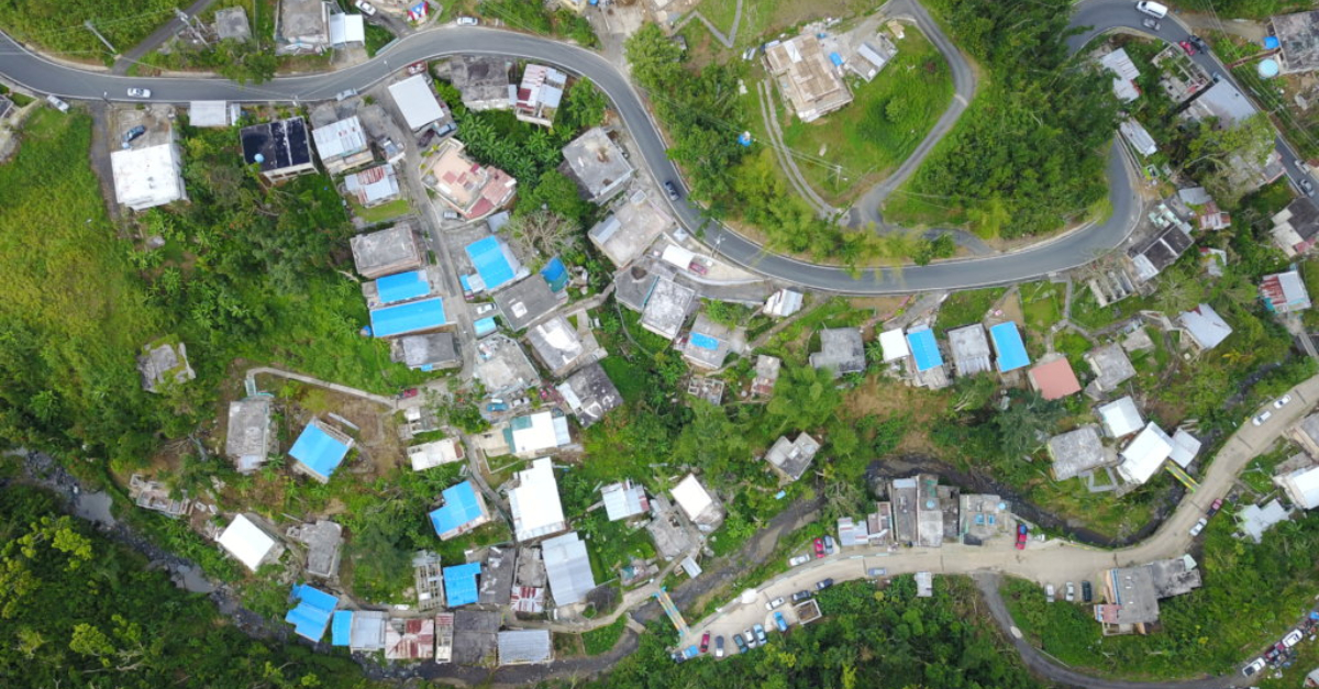 An aerial view of houses, many with blue roofs, and winding roads. Green grass and trees surround the houses and roads.