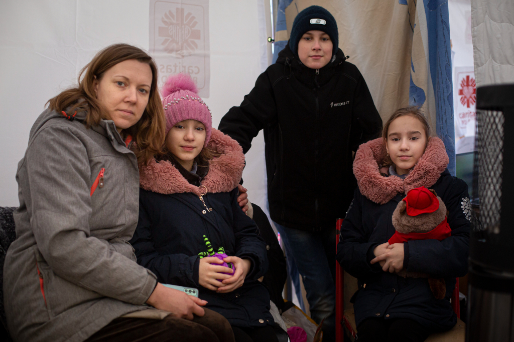 A woman sits with her three children inside a refugee tent. All three are wearing winter jackets. Nonprofits helping Ukraine.