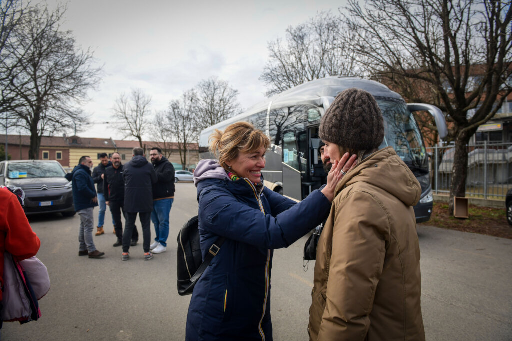 A woman wearing a blue jacket and backpack smiles and puts her hands on the face of a woman wearing a brown jacket and hat. Other people stand in the background. Nonprofits helping Ukraine.