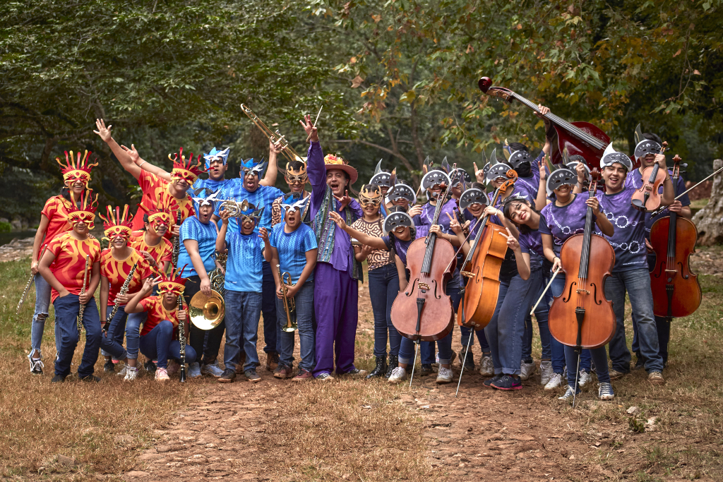 Large group of children and teachers dressed in festival colors and masks, and holding instruments, wave their hands and smile at the camera - photos of 2022