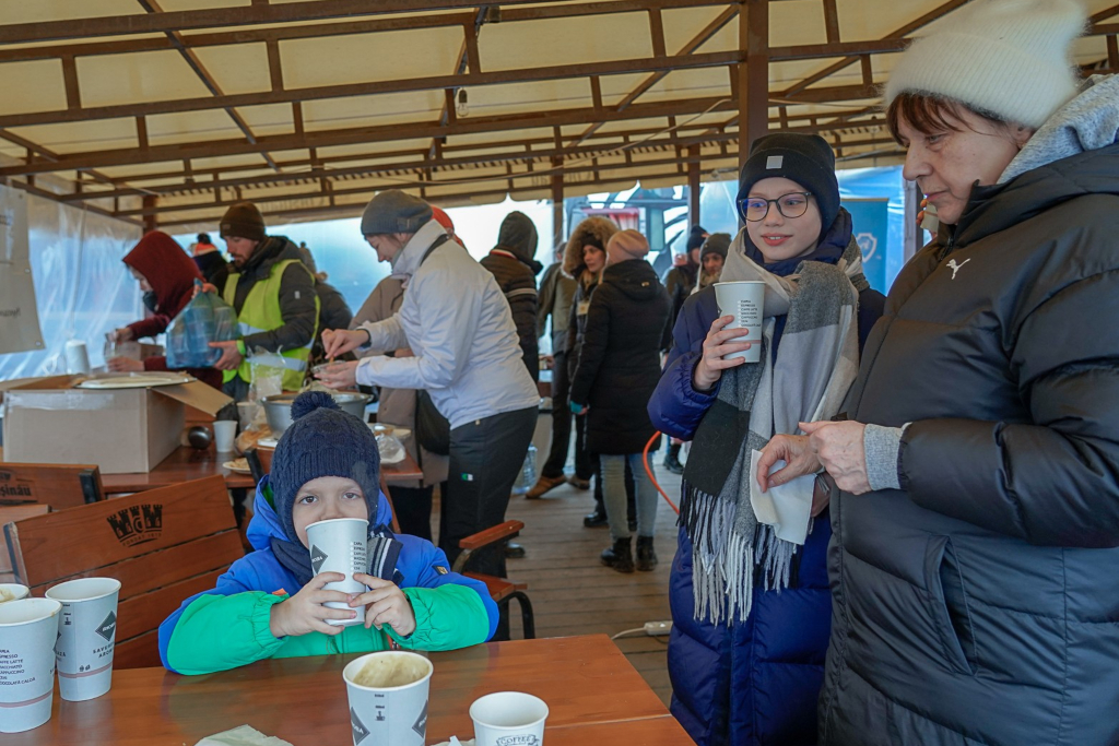 responding to crisis, many people, dressed in warm clothes, gather under a canopy getting food and drinking hot beverages