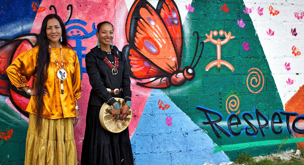 Two women stand in front of a mural