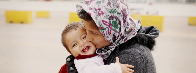 A photo of a woman in a headscarf kissing a smiling toddler on the cheek