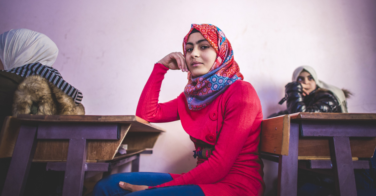 A young woman in a pink shirt sits at a school desk. Syrian refugee crisis facts