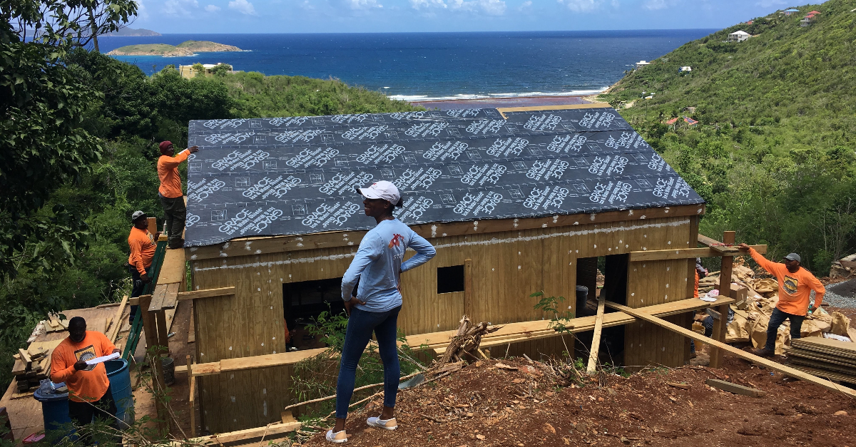 A person stands on a hill overlooking a house under construction and a crew of workers in orange shirts. The blue sea is behind the house in the distance.