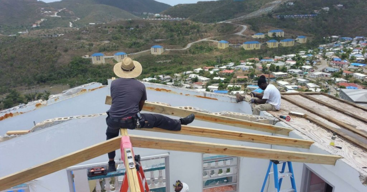 A construction worker wearing a wide-brimmed hat sits on top of a partially constructed roof overlooking St. Thomas. Another worker sits on the edge of the frame.