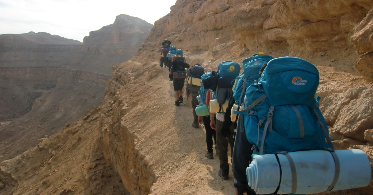 A line of backpackers with blue packs hike near the edge of a cliff