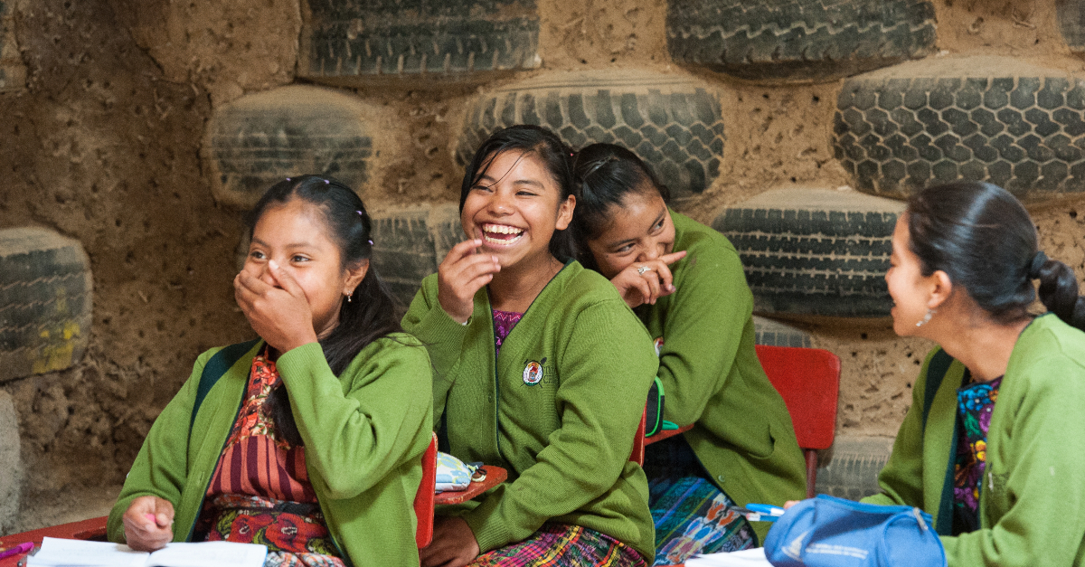 Four female students in green sweaters laugh with each other in a classroom made from tires and mud.