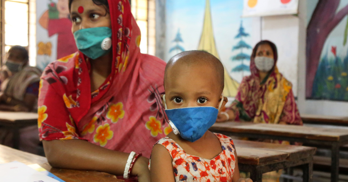 A child in a blue mask looks straight ahead and stands in front of a woman wearing a blue mask and pink saree as she looks to the side.