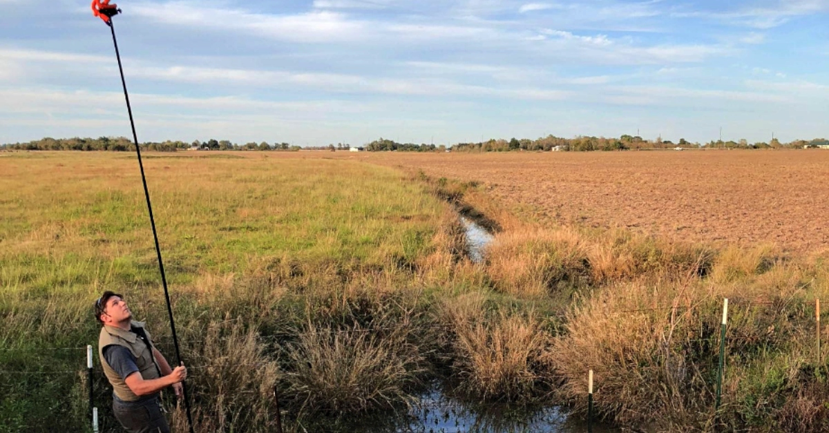 A man in a green vest stands in a wetland holding a long device in his hands to monitor the area. A grassy field is behind him.