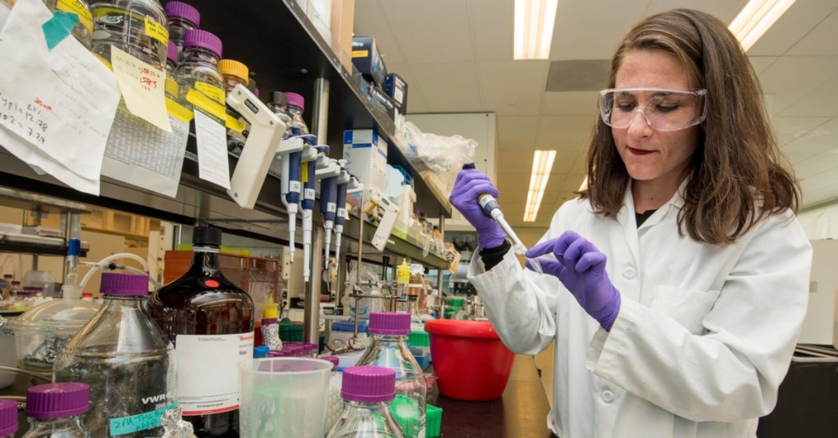 A woman in a white lab coat and goggles puts liquid from a syringe into a test tube