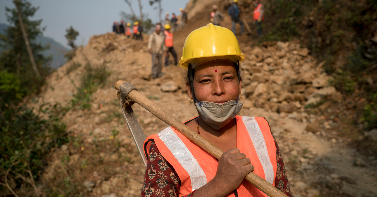 A woman in a yellow hard hat and an orange safety vest stands with a hoe on her shoulder. She has a face mask pulled down on her chin. Other workers are out of focus in the background.