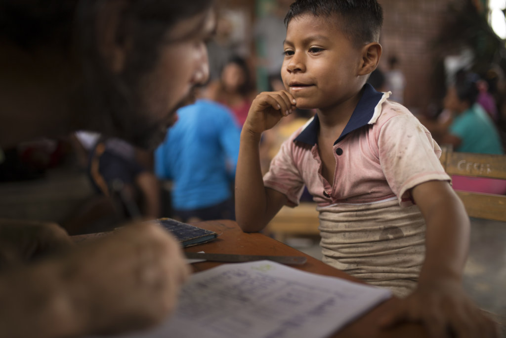 A young boy in a faded red shirt with a blue collar listens with his right hand on his chin. His teacher looks at him in the foreground.