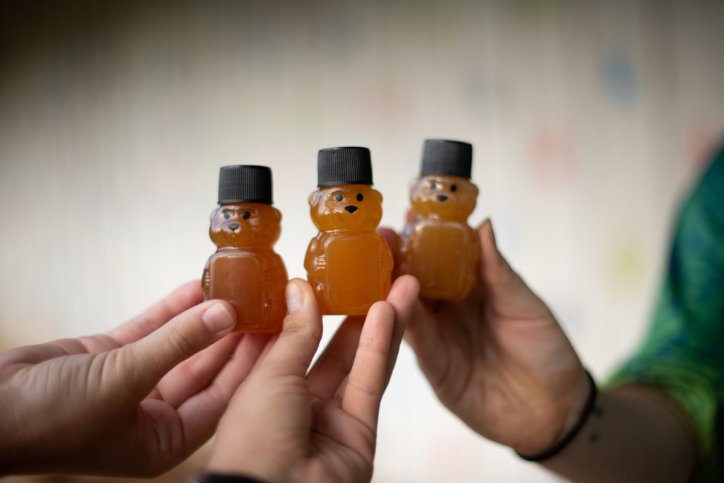 Three hands hold up small bear-shaped bottles of stingless bee honey.