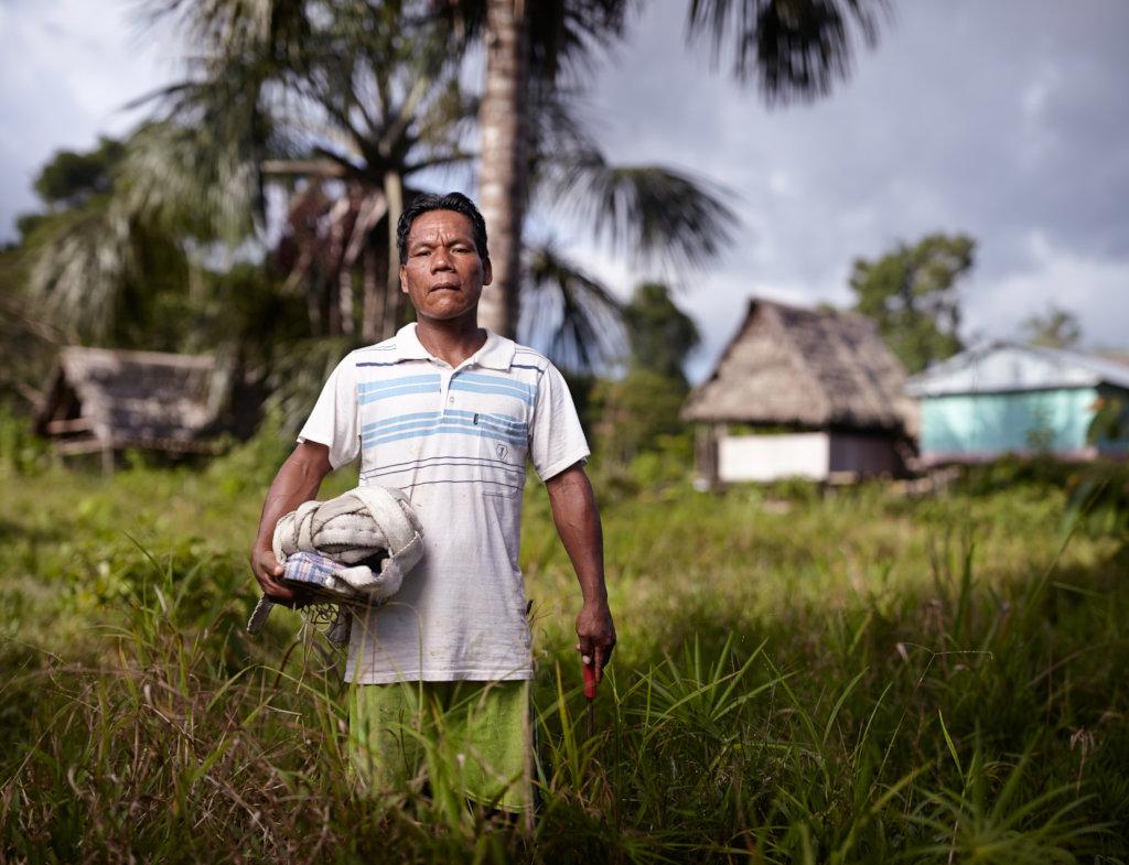 A man in a white polo shirt with blue stripes holds a machete in his left hand and thick rope in his right. Palm trees and houses with thatched roofs are in the background.