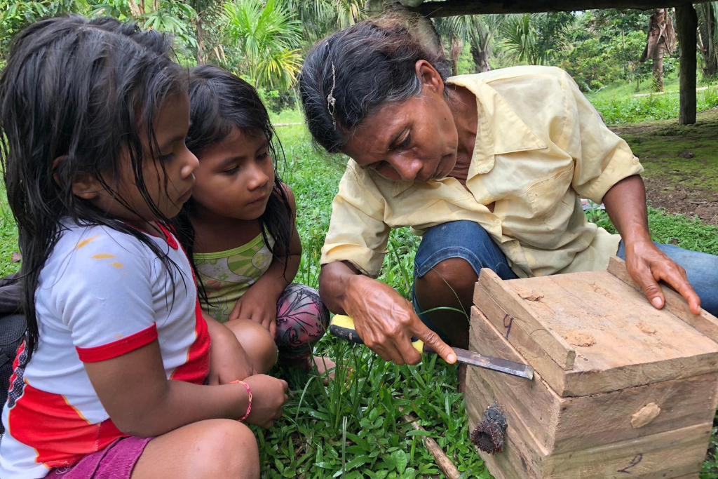 A grandmother in a yellow shirt opens a beehive box with a chisel. Her two young granddaughters watch.