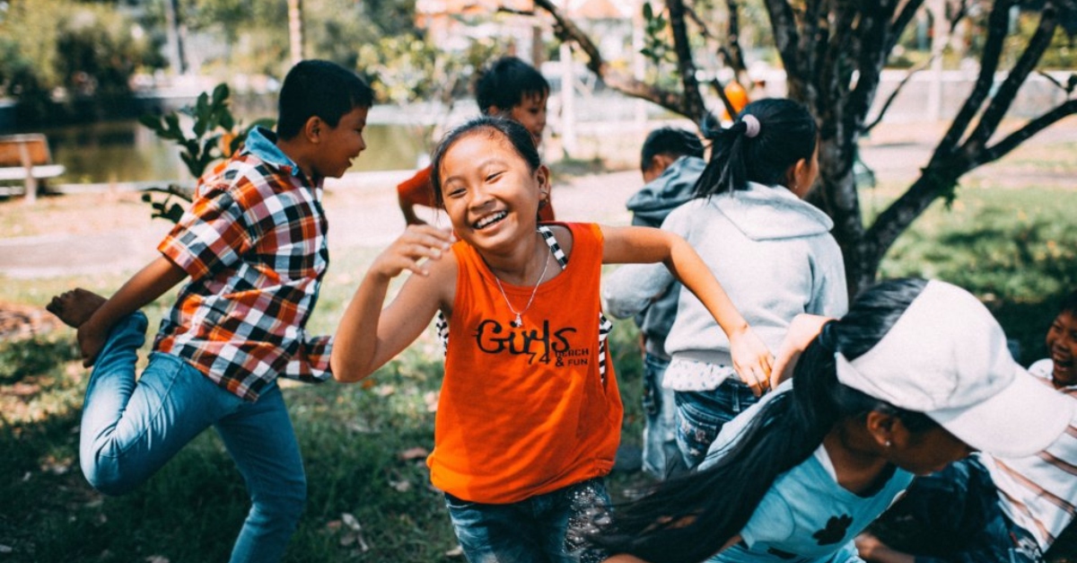 A girl in an orange tanktop smiles as she plays tag with other children.