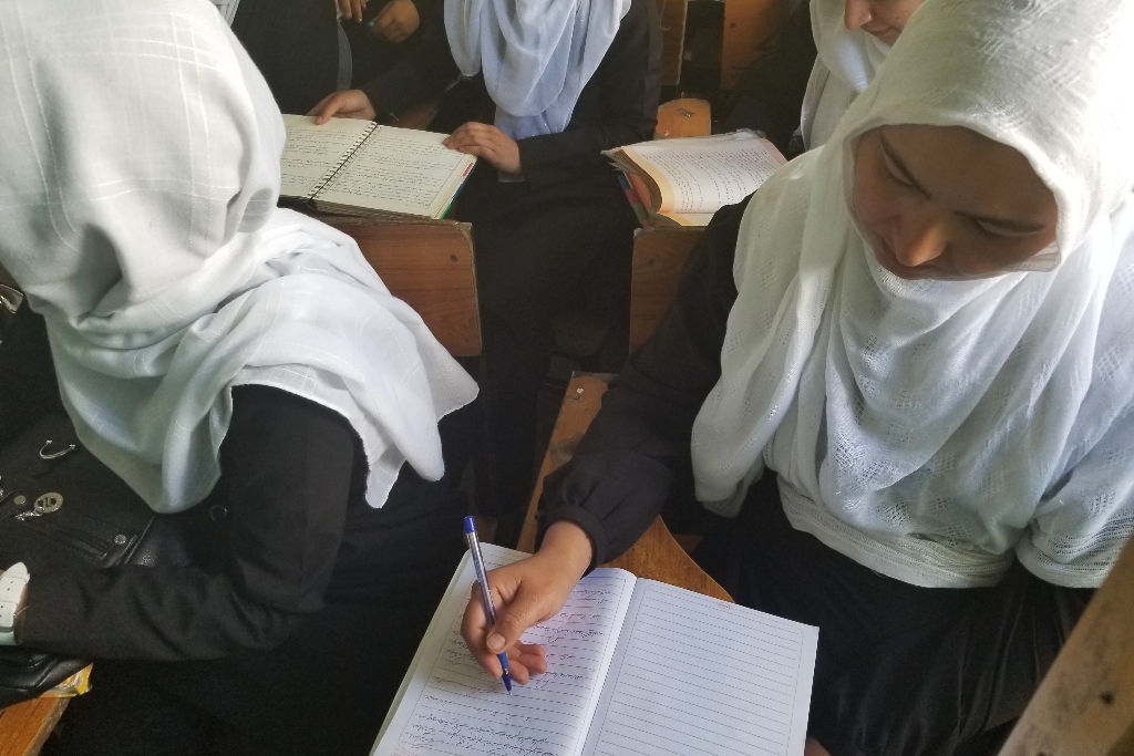 A girl in a white hijab writes in a notebook at a school desk. Other students are around her. Refugee stories