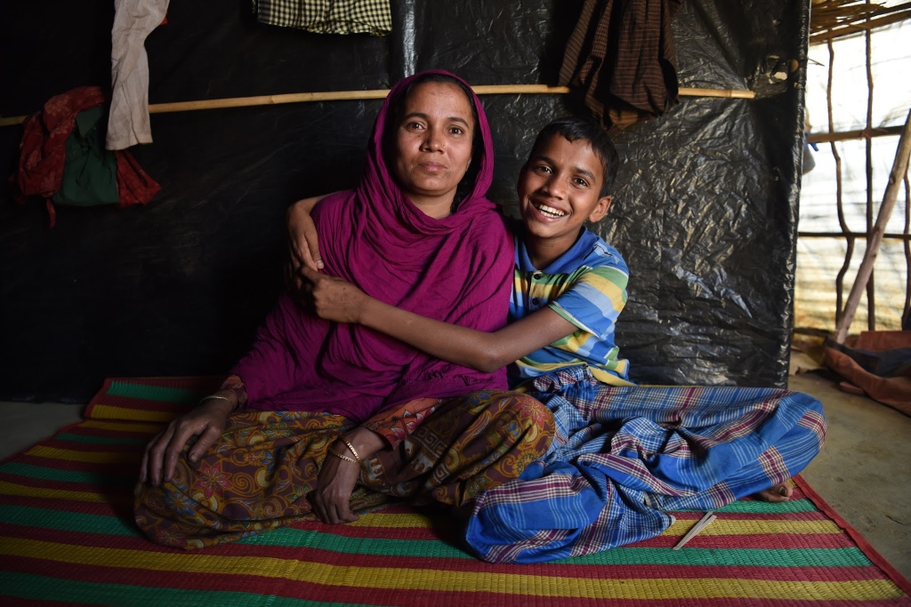 A boy in a blue and yellow t-shirt embraces his mother who is wearing a purple head covering. They sit on a carpet inside a tent