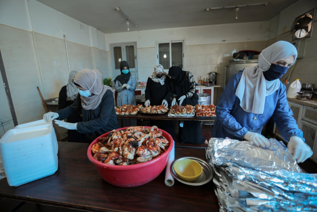 Women in gloves and hijabs prepare to package chicken in aluminum foil for delivery