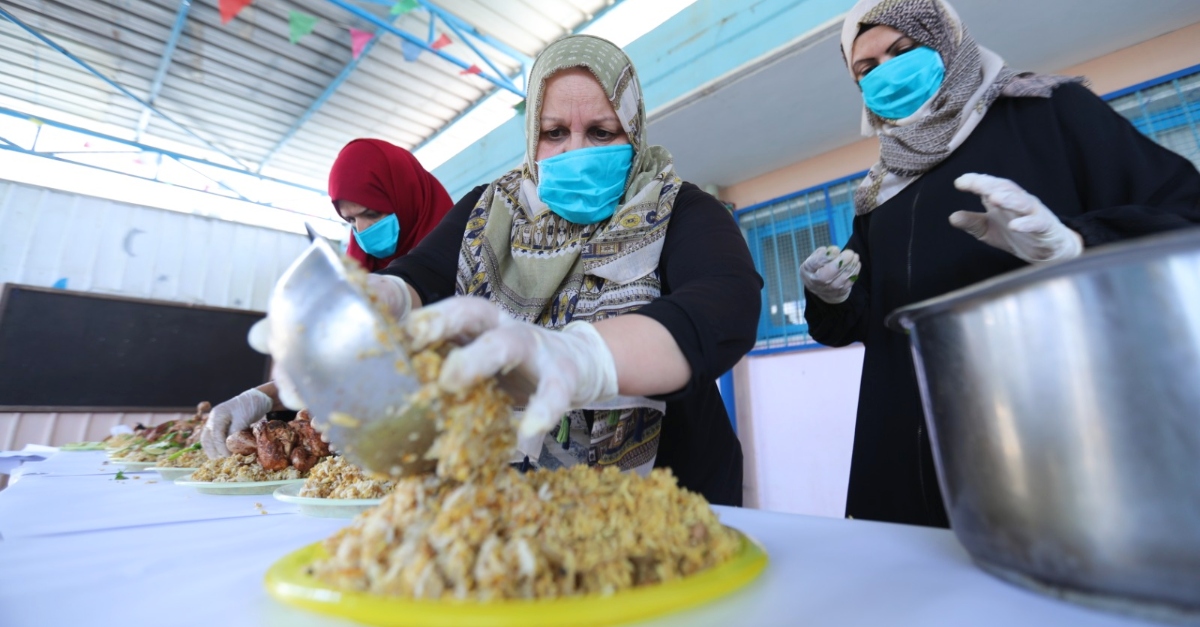 Mrs. Najah in a blue mask and green patterned hijab serves rice onto a place. Two volunteer cooks in blue masks stand next to her.