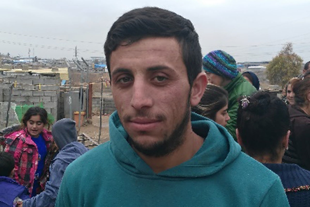 A man in a teal sweatshirt smiles faintly. People fill the background