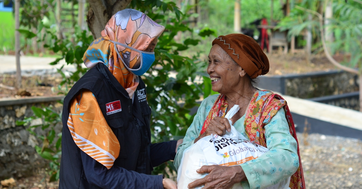 A woman in a headscarf and a blue mask gives a bag with emergency food supplies, funded by Facebook's crisis response program, to a woman in a brown hat who is smiling.