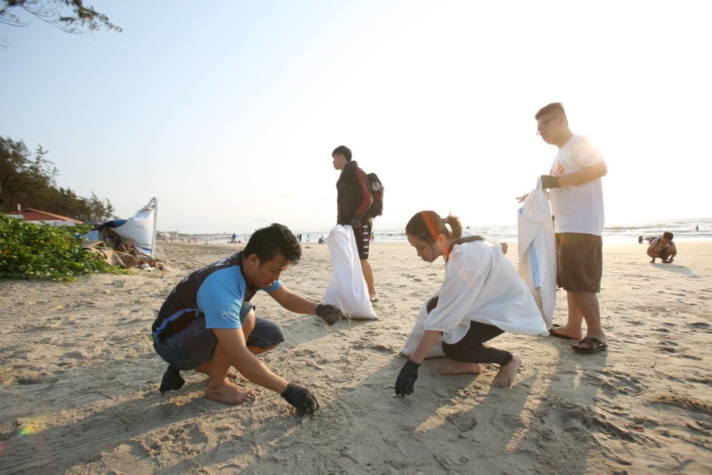 Young people pick up trash on a beach with the sun shining behind them