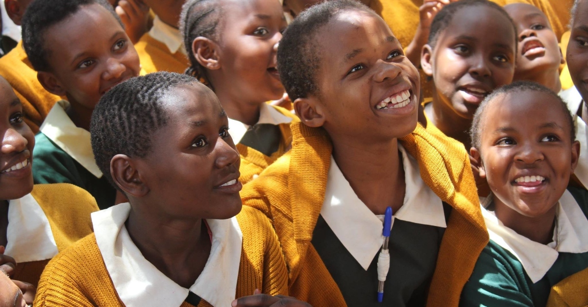 A group of children in green school uniforms and yellow cardigans look up, smiling