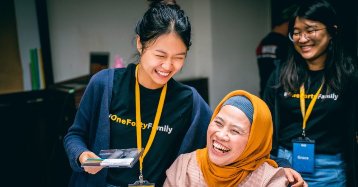 A woman wearing a T-shirt and lanyard and holding markers laughs with another woman in a yellow hijab. In the background, a colleague wears a matching lanyard and T-shirt.