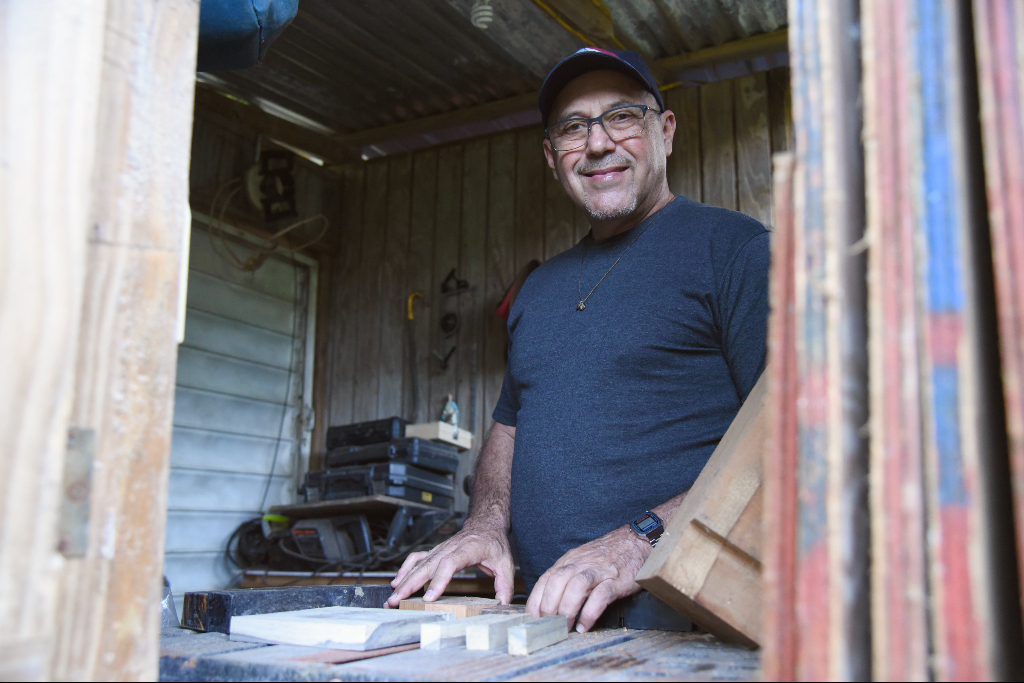 A man in a blue t-shirt and hat touches blocks of wood on a table saw in front of him.