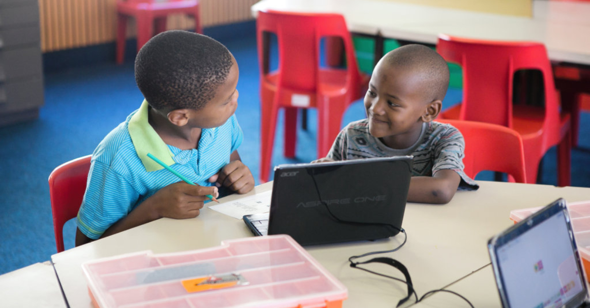 Two young boys sit at a classroom desk behind a laptop. Red chairs and a table are in the background.