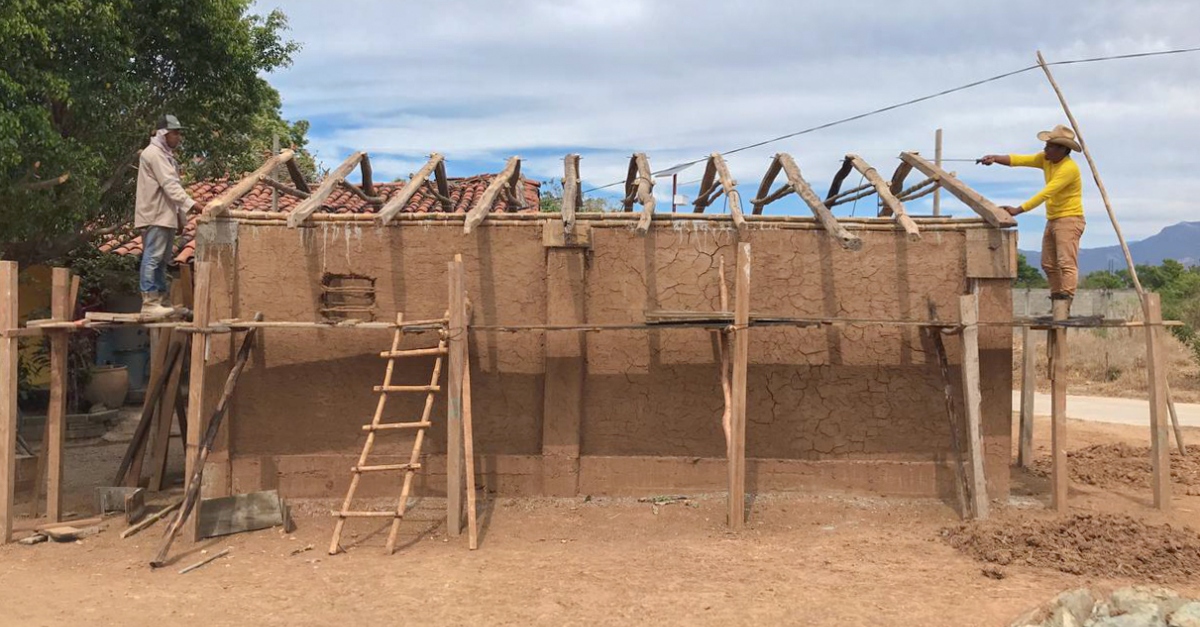 Mexico earthquake before and after. Two men stand on wooden scaffolding at either end of the frame of a house they are building with wood and a mud mixture.