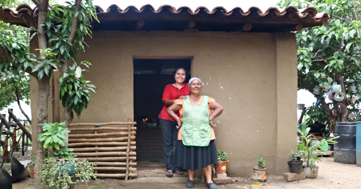A woman in a green apron and a woman in a red shirt stand in the doorway of a brown house. Potted plants and trees surround it.